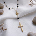 A gold cross pendant necklace with small black beads and a round gold disc charm, styled on draped white fabric with decorative stones and dried flowers.