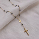 Rosario black beaded cross pendant close up- A delicate gold chain necklace with small black beads, a small round gold medallion, and a cross pendant, displayed on a textured off-white fabric background.