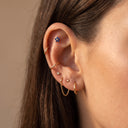 Sapphire Flora Flower Piercing Stud. Close-up of a person's ear showcasing multiple gold ear piercings, including a small blue flower stud, a curved gold hoop, a huggie, and chain details, against a warm beige background.