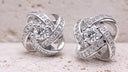 Pair of silver knot earrings with clear gemstones on a textured beige surface