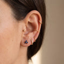 Sapphire Trio Piercing Stud- Close-up of a person's ear showcasing multiple gold ear piercings and earrings, including a hoop in the lobe, a chain in the cartilage, a small stud, and a cluster of blue stones, against a soft beige background.