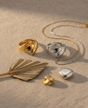 A styled flat-lay of jewelry on a textured beige fabric background, featuring a gold wrap ring, a silver wrap ring, a gold chain necklace, a gold fan-shaped leaf, a small gold chunk, and a polished silver stone.